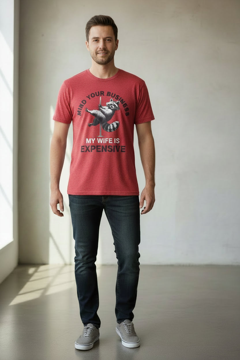 A red heather graphic t-shirt featuring a raccoon lifting a barbell with the text 'MIND YOUR BUSINESS' and 'MY WIFE IS EXPENSIVE' printed on the front.