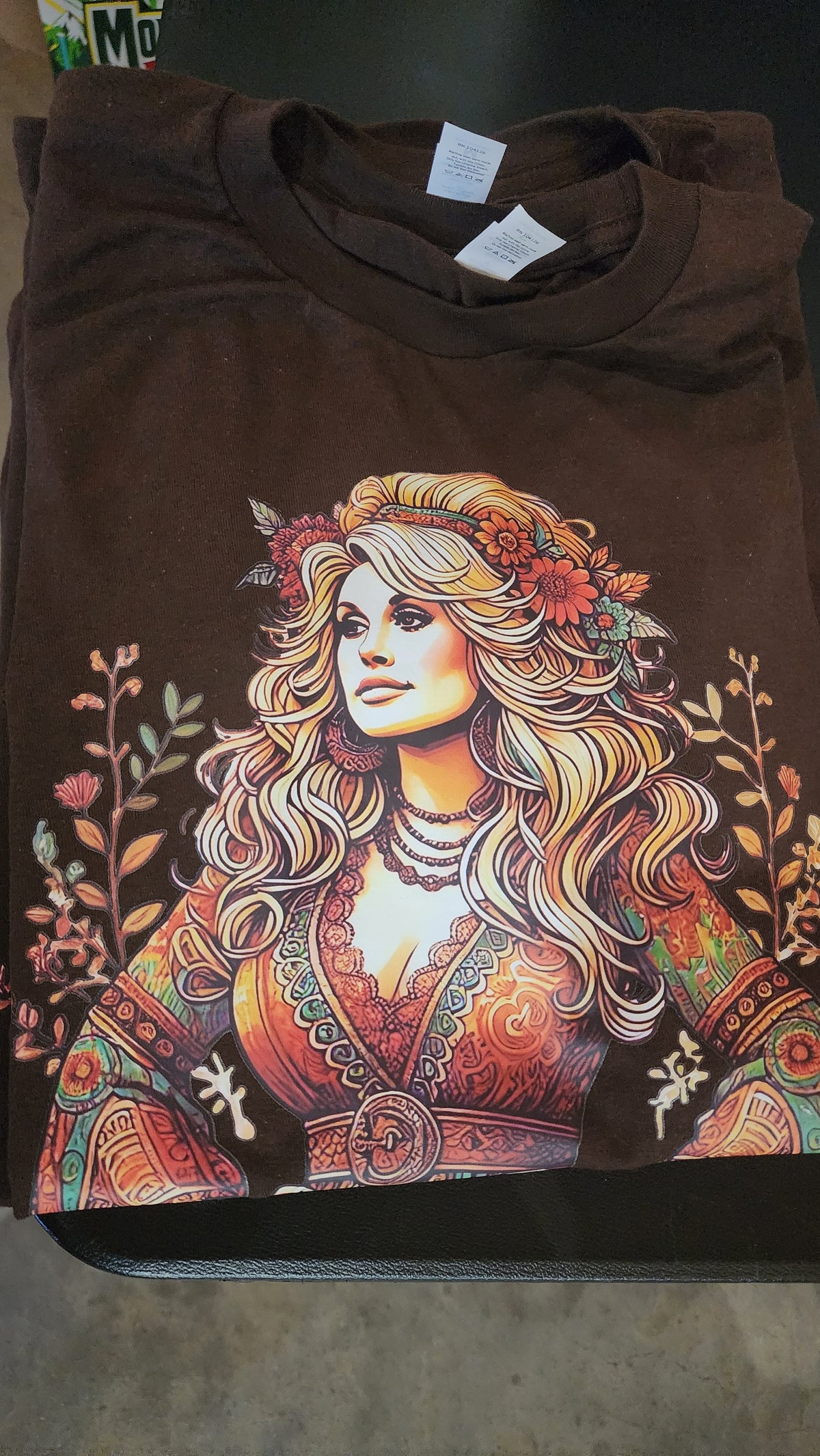 A folded brown t-shirt featuring a large graphic print of a woman with long blonde wavy hair, wearing a bohemian-style patterned dress and floral headpiece, displayed on a dark surface.
